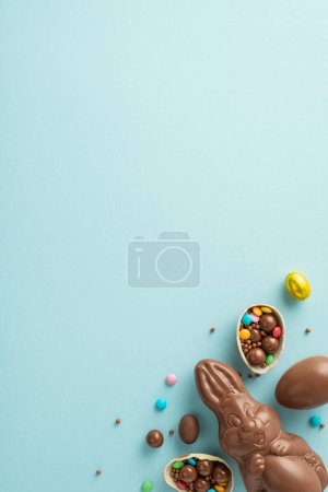 Inviting Easter compilation scheme. Vertical top down view of chocolate egg broken apart, filled with colorful candies, chocolate bunny, sprinkles on a pastel blue backdrop, with space for words