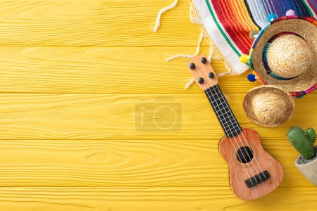 Cinco de Mayo decor: top view of sombreros, guitar, and a cactus. Colorful serape on a yellow wooden desk. Ideal for text placement