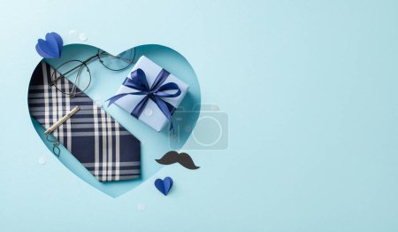 Foto de Di "Feliz Día del Padre" con sofisticación. Vista superior de la corbata con clip, gafas, caja de regalo y más, enmarcada en un recorte en forma de corazón en azul pastel. Ideal para saludos o promociones - Imagen libre de derechos