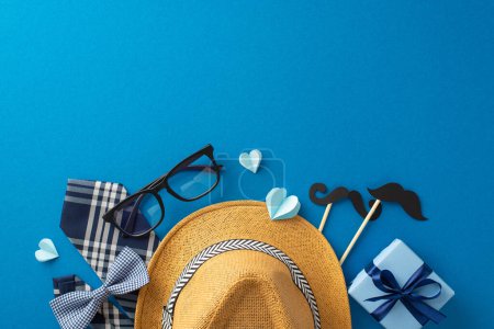 Creative flat lay of Father's Day gift items including a stylish straw hat, glasses, bow tie, and mustache props against a bold blue background