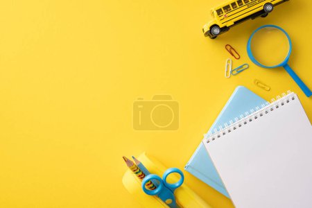 Flat lay of back to school supplies including a notebook, crayons, scissors, and a toy school bus on a bright yellow background