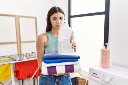 Foto de Mujer hispana joven haciendo la colada sosteniendo la botella de detergente y doblando la ropa deprimida y preocupada por la angustia, llorando enojada y asustada. expresión triste. - Imagen libre de derechos
