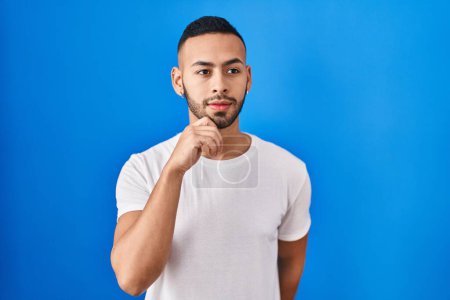 Photo for Young hispanic man standing over blue background with hand on chin thinking about question, pensive expression. smiling and thoughtful face. doubt concept. - Royalty Free Image