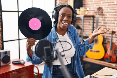 Foto de Beautiful black woman holding vinyl record at music studio celebrating achievement with happy smile and winner expression with raised hand - Imagen libre de derechos