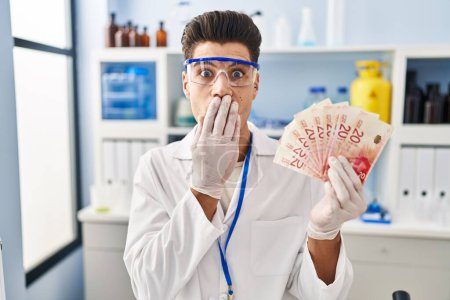 Photo for Young hispanic man working at scientist laboratory holding shekels covering mouth with hand, shocked and afraid for mistake. surprised expression - Royalty Free Image