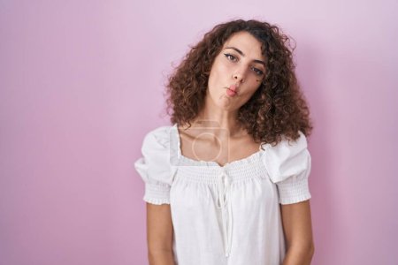 Foto de Hispanic woman with curly hair standing over pink background making fish face with lips, crazy and comical gesture. funny expression. - Imagen libre de derechos