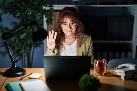 Téléchargez les photos : Moyen Age femme hispanique travaillant à l'aide d'un ordinateur portable la nuit montrant et pointant vers le haut avec les doigts numéro quatre tout en souriant confiant et heureux. - en image libre de droit