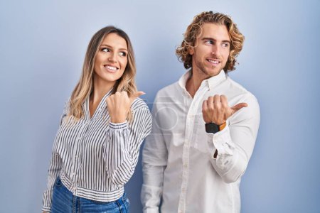 Foto de Young couple standing over blue background smiling with happy face looking and pointing to the side with thumb up. - Imagen libre de derechos