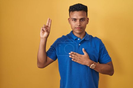 Foto de Joven hispano de pie sobre fondo amarillo sonriendo jurando con la mano en el pecho y los dedos hacia arriba, haciendo un juramento de lealtad promesa - Imagen libre de derechos
