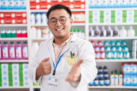 Photo pour Jeune homme chinois travaillant à la pharmacie pharmacie souriant joyeux offrant la main de la paume donnant assistance et acceptation. - image libre de droit
