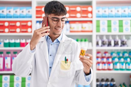 Téléchargez les photos : Jeune homme hispanique pharmacien tenant bouteille de pilules parler sur smartphone à la pharmacie - en image libre de droit