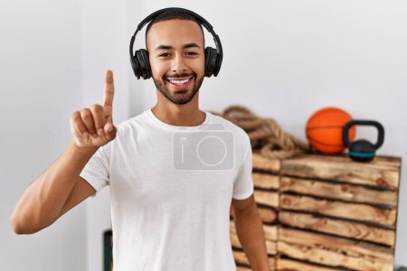 Téléchargez les photos : African american man listening to music using headphones at the gym showing and pointing up with finger number one while smiling confident and happy. - en image libre de droit