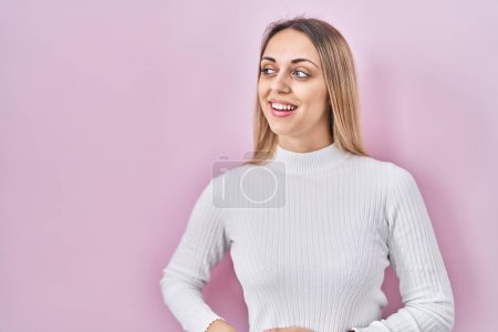 Photo for Young blonde woman wearing white sweater over pink background looking away to side with smile on face, natural expression. laughing confident. - Royalty Free Image