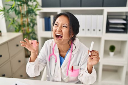 Téléchargez les photos : Jeune médecin asiatique femme tenant la coupe menstruelle et tampon en colère et fou hurlant frustré et furieux, criant de colère. rage et concept agressif. - en image libre de droit