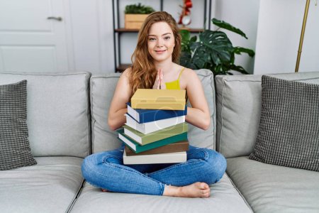 Photo for Young caucasian woman sitting on the sofa with books at home praying with hands together asking for forgiveness smiling confident. - Royalty Free Image