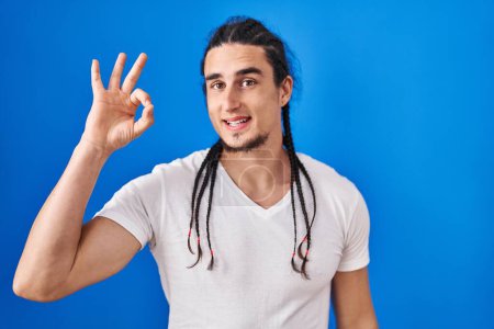 Foto de Hombre hispano con el pelo largo de pie sobre fondo azul sonriendo positivo haciendo signo de ok con la mano y los dedos. expresión exitosa. - Imagen libre de derechos