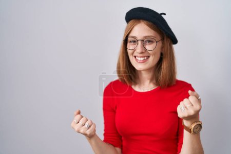 Photo for Young redhead woman standing wearing glasses and beret very happy and excited doing winner gesture with arms raised, smiling and screaming for success. celebration concept. - Royalty Free Image