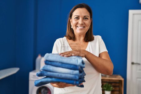 Téléchargez les photos : Middle age hispanic woman smiling confident holding folded jeans at laundry room - en image libre de droit