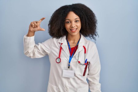 Téléchargez les photos : Jeune femme afro-américaine portant l'uniforme de médecin et stéthoscope geste souriant et confiant avec la main faisant signe de petite taille avec les doigts regardant et la caméra. concept de mesure. - en image libre de droit
