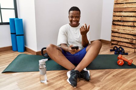 Foto de Joven hombre africano sentado en la alfombra de entrenamiento en el gimnasio usando el teléfono inteligente señalización ven aquí gesto con la mano invitando a la bienvenida feliz y sonriente - Imagen libre de derechos