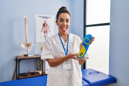 Foto de Mujer hispana joven sosteniendo plantilla de zapato en clínica de fisioterapia sacando la lengua feliz con expresión divertida. - Imagen libre de derechos