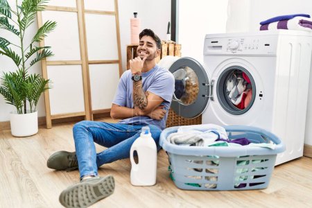Photo for Young hispanic man putting dirty laundry into washing machine with hand on chin thinking about question, pensive expression. smiling and thoughtful face. doubt concept. - Royalty Free Image