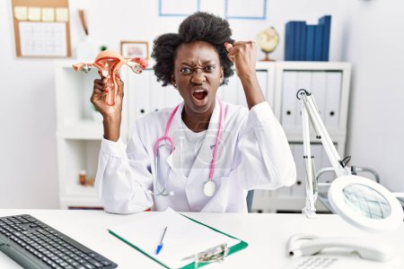 Téléchargez les photos : African doctor woman holding anatomical model of female genital organ at medical clinic annoyed and frustrated shouting with anger, yelling crazy with anger and hand raised - en image libre de droit