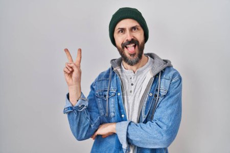 Photo for Young hispanic man with tattoos wearing wool cap smiling with happy face winking at the camera doing victory sign. number two. - Royalty Free Image