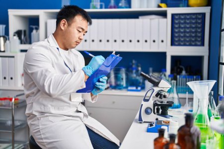 Téléchargez les photos : Young chinese man wearing scientist uniform writing on clipboard at laboratory - en image libre de droit