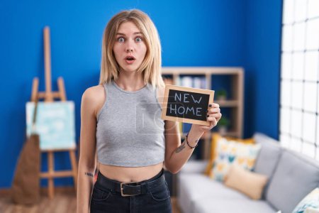 Photo for Blonde caucasian woman holding blackboard with new home text scared and amazed with open mouth for surprise, disbelief face - Royalty Free Image