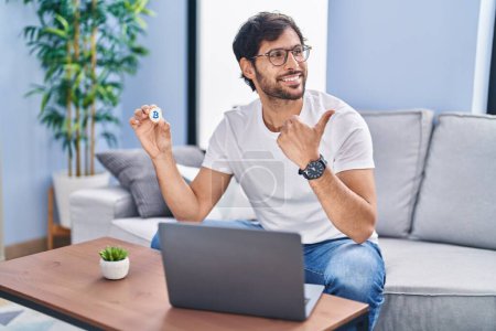 Photo for Handsome latin man holding virtual currency bitcoin using laptop pointing thumb up to the side smiling happy with open mouth - Royalty Free Image