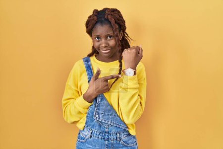 Photo for African woman standing over yellow background in hurry pointing to watch time, impatience, looking at the camera with relaxed expression - Royalty Free Image