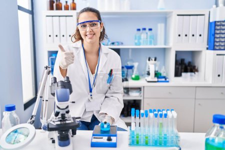 Foto de Young hispanic woman working at scientist laboratory doing happy thumbs up gesture with hand. approving expression looking at the camera showing success. - Imagen libre de derechos