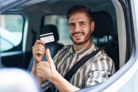 Téléchargez les photos : Homme hispanique avec barbe voiture de conduite tenant carte de crédit souriant heureux et positif, pouce levé faire excellent et signe d'approbation - en image libre de droit