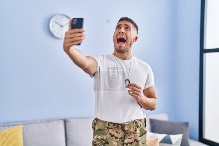 Foto de Joven hombre hispano usando uniforme camuflaje del ejército tomando selfie en casa enojado y loco gritando frustrado y furioso, gritando con ira mirando hacia arriba. - Imagen libre de derechos