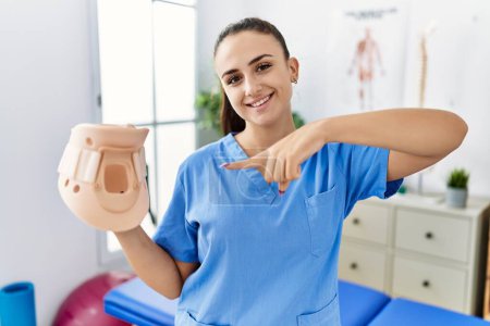 Téléchargez les photos : Jeune physiothérapeute femme tenant col cervical à la clinique médicale souriant heureux pointant avec la main et le doigt - en image libre de droit