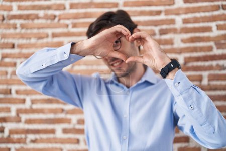 Téléchargez les photos : Jeune homme hispanique debout sur fond de mur de briques faisant forme de coeur avec la main et les doigts souriant en regardant à travers le signe - en image libre de droit