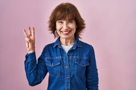 Photo for Middle age woman standing over pink background showing and pointing up with fingers number three while smiling confident and happy. - Royalty Free Image