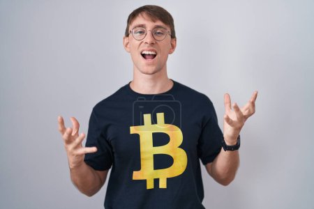 Photo for Caucasian blond man wearing bitcoin t shirt crazy and mad shouting and yelling with aggressive expression and arms raised. frustration concept. - Royalty Free Image