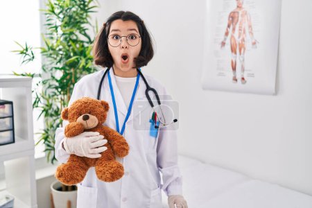 Foto de Joven doctora hispana sosteniendo oso de peluche asustada y sorprendida con la boca abierta para sorpresa, cara de incredulidad - Imagen libre de derechos