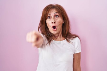 Photo for Middle age woman standing over pink background pointing with finger surprised ahead, open mouth amazed expression, something on the front - Royalty Free Image