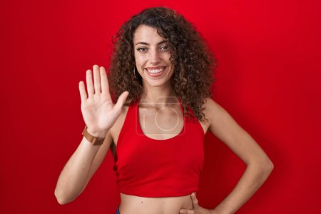 Photo for Hispanic woman with curly hair standing over red background waiving saying hello happy and smiling, friendly welcome gesture - Royalty Free Image