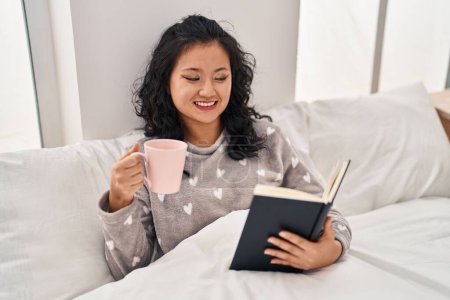 Téléchargez les photos : Jeune femme chinoise dînant café et livre de lecture assis sur le lit dans la chambre - en image libre de droit