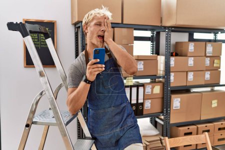 Téléchargez les photos : Young blond man using smartphone working at storehouse covering one eye with hand, confident smile on face and surprise emotion. - en image libre de droit