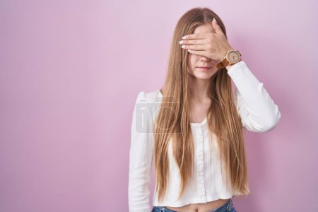 Téléchargez les photos : Young caucasian woman standing over pink background covering eyes with hand, looking serious and sad. sightless, hiding and rejection concept - en image libre de droit