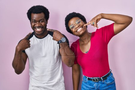 Téléchargez les photos : Jeune couple afro-américain debout sur fond rose souriant et joyeux montrant et pointant du doigt les dents et la bouche. concept de santé dentaire. - en image libre de droit