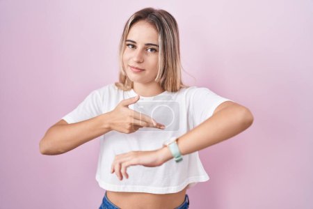 Foto de Young blonde woman standing over pink background in hurry pointing to watch time, impatience, upset and angry for deadline delay - Imagen libre de derechos