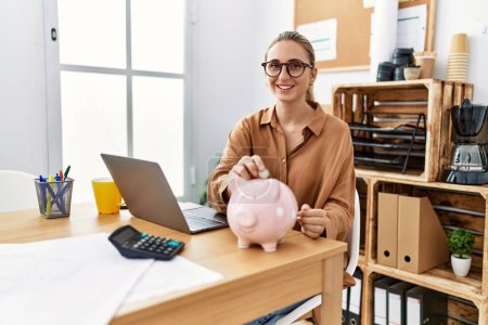 Photo for Young blonde woman at the office putting coin into piggy bank looking positive and happy standing and smiling with a confident smile showing teeth - Royalty Free Image