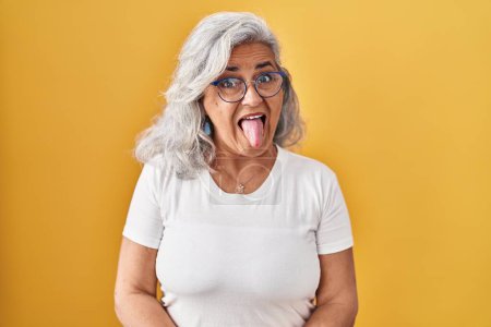 Foto de Mujer de mediana edad con el pelo gris de pie sobre el fondo amarillo sacando la lengua feliz con expresión divertida. concepto de emoción. - Imagen libre de derechos