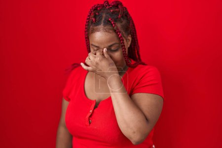 Foto de African american woman with braided hair standing over red background tired rubbing nose and eyes feeling fatigue and headache. stress and frustration concept. - Imagen libre de derechos